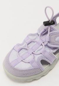Violeta sānce ar acu tīklu augšdaļu, elastīgām šņorēm, mīkstiem siksnām un teksturētu zoli. Ietver regulējamu fiksatoru pielāgošanai.