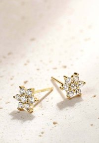 Pendientes de aro dorados en forma de flores, adornados con múltiples piedras preciosas transparentes en un diseño agrupado, sobre un fondo texturizado claro.