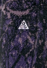 Fleece jacket with a black and purple camouflage pattern. Features a triangular silver logo and a visible zipper detail. Soft texture.