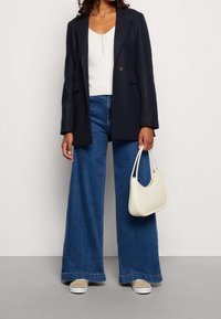 Marineblazer, weißes geripptes Top mit Knöpfen, weit geschnittene blaue Jeans, beige Sneakers und weiße Handtasche. Kombination von Texturen und Farben.