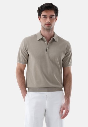 Man wearing beige short-sleeve polo shirt with white trim and white pants, standing against a plain light background.