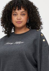 Kolgrå sweatshirt med broderad "Tommy Hilfiger" logotyp; har guldknappar på axlarna och en avslappnad passform.