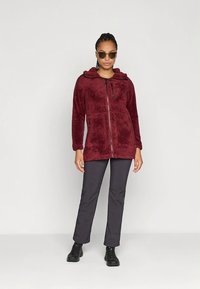 Vinrød fleecejakke med glidelås foran, hette og to sidelommer, kombinert med mørkegrå friluftsbukser og svarte støvler. Glatt tekstur.