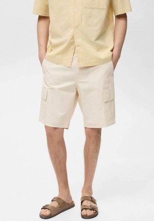 Beige Cargo-Shorts mit zwei seitlichen Taschen, gerade geschnitten, aus leichtem Stoff. Kombiniert mit einem hellgelben Kurzarmhemd.