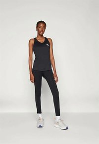 Kvinna som står iförd en svart ärmlös träningsoberå, svarta leggings, vita strumpor och vita löparskor med orangea och blå accenter.