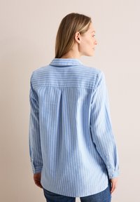 Camicia a maniche lunghe azzurro chiaro con strisce bianche verticali, colletto e polsini con bottoni, hem arcuato e dettaglio nella parte posteriore.
