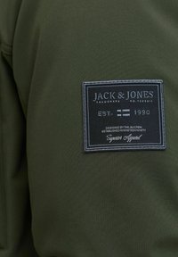 Groen textiel kledingstuk met een zwart stoffen label met witte tekst, inclusief merknaam "JACK & JONES," oprichtingsdatum en ontwerpinformatie.