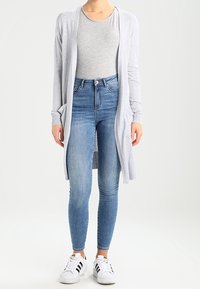 Cardigan gris claro hecho de un material de punto suave, llevado sobre una camiseta gris. Combinado con jeans azules de cintura alta y zapatillas blancas.