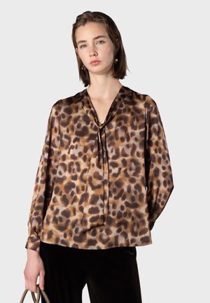 Kvinde iført en brun og sort leopardmønstret bluse med bindebånd ved halsen, der holder en håndtaske, mod en ensartet lys baggrund.