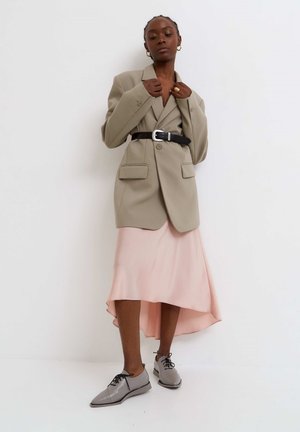 Blazer beige oversize con due tasche anteriori, cintato in vita con una cintura nera. Indossato sopra un vestito in satin rosa pastello e scarpe grigie.