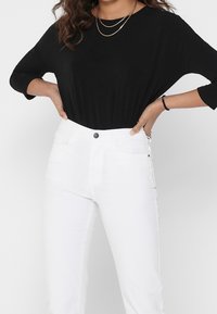 Schwarzes Langarmoberteil kombiniert mit hoch taillierten weißen Jeans. Das Oberteil hat einen lässigen Schnitt und die Jeans verfügen über einen Knopfverschluss und Taschen.