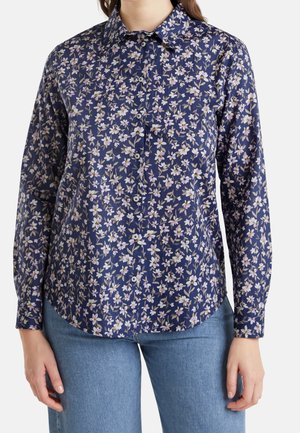 Mujer con una camisa azul marino abotonada con un pequeño estampado floral blanco y lila claro y vaqueros azul claro.