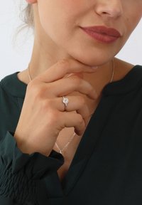 Silberner Ring mit einem runden, funkelnden Mittelpunkt, auf einem schmalen Band gesetzt. Die Hand des Models ruht auf ihrem Kinn und sie trägt ein dunkelgrünes Oberteil.