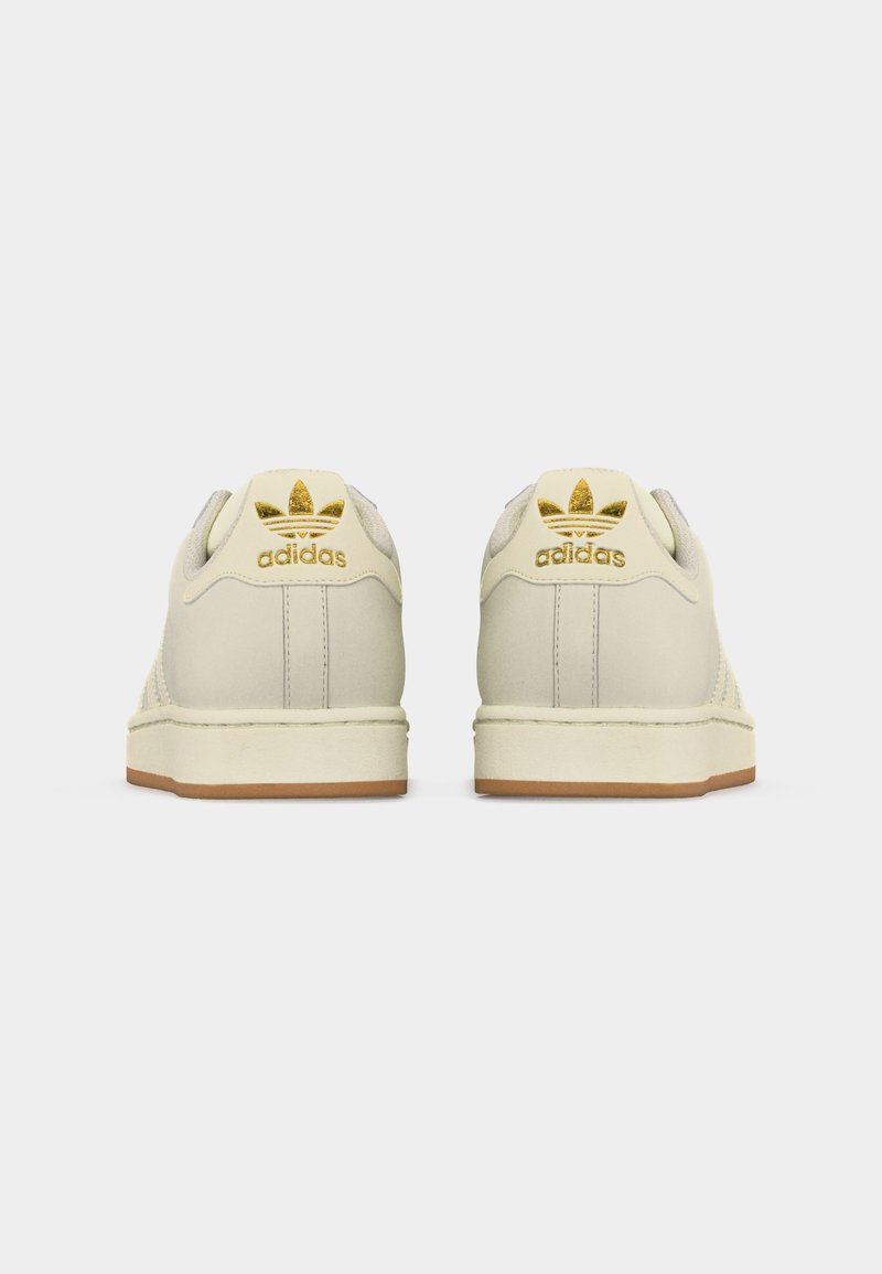 Paire de baskets Adidas couleur crème vues de dos, avec logo Adidas doré et semelles couleur gomme sur un fond blanc.