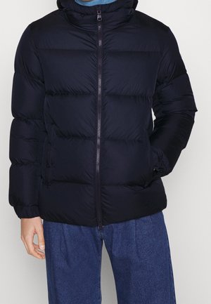 Mężczyzna w czarnej puchowej kurtce z przednim zamkiem błyskawicznym, ręce częściowo w kieszeniach, ubrany w luźne niebieskie dżinsy na jednolitym tle.