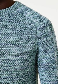 Close-up van een gebreide ronde halstrui in blauw, groen en grijstinten met gestructureerde steekpatronen op de schouder en mouw.