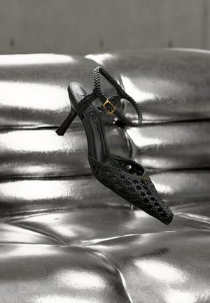 Čierna kožená sandála na vysokom podpätku s členkovým remienkom a zlatou prackou, vystavená nad lesklým strieborným čalúneným povrchom.