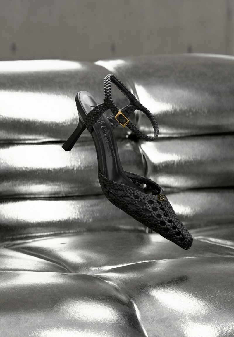 Μαύρο υφαντό δερμάτινο σανδάλι με ψηλό τακούνι και λουράκι στον αστράγαλο με χρυσή αγκράφα, τοποθετημένο πάνω σε μια λαμπερή ασημένια επιφάνεια με καπιτονέ λεπτομέρειες.