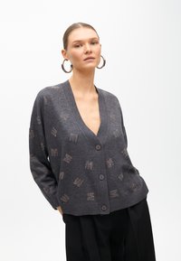 Sivý cardigan s výstrihom do V, zapínaním na gombíky a dekoratívnym študovaným vzorom. Vyrobený z mäkkého materiálu s dlhými rukávmi a voľným strihom.