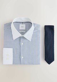 Chemise à rayures bleu et blanc avec col et poignets blancs, posée à côté d'une cravate bleu foncé texturée sur une surface blanche.