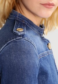 Tummansininen farkkutakki, muotoiltu malli, jossa on näkyvät kultaiset napit olkapäissä ja hienovaraisia ompeluyksityiskohtia. Pintakudos näyttää sileältä.