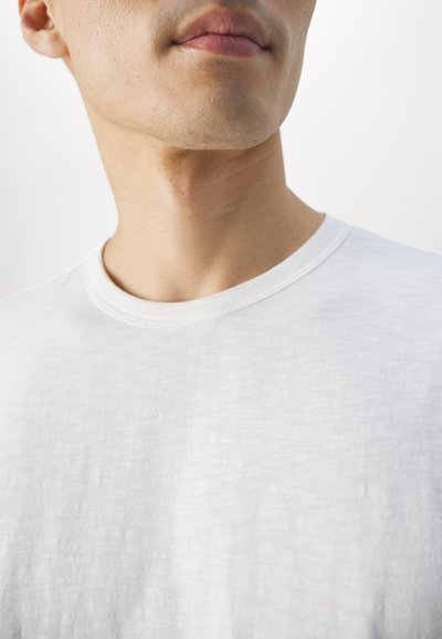 T-shirt bianco a girocollo realizzato in tessuto leggero, con superficie testurizzata e scollatura liscia, visibile sopra la clavicola.