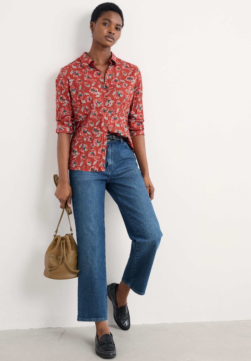 Camicia a bottone rossa con fantasie floreali, jeans blu a gamba dritta, mocassini neri e borsa a secchiello beige. Presenta maniche arrotolate e tessuto texturizzato.