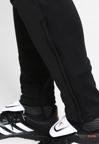 Untere Beine in schwarzen, schmal zulaufenden Hosen mit seitlichen Reißverschlüssen und schwarzen Adidas Copa Fußballschuhen mit weißen Zungenklappen und schwarzen Schnürsenkeln.