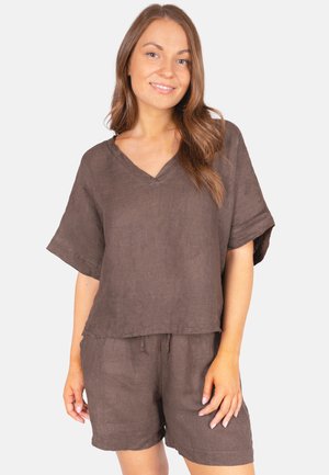 Jeune femme aux longs cheveux bruns portant un haut marron ample à manches courtes et un short assorti, souriant avec un fond neutre.