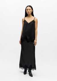 Camisole en satin noir avec bordure en dentelle associée à une jupe longue assortie, présentant une texture lisse et un design minimaliste. Des bottines noires complètent le look.