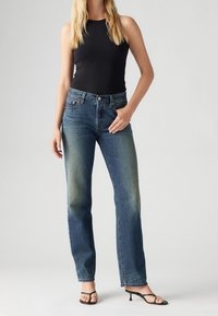 Džínové kalhoty s vysokým pasem a rovnými nohavicemi v modré barvě, s vybledlými stehnami, pět kapsami a surovým lemem. V kombinaci s černým topem bez rukávů a černými sandály.