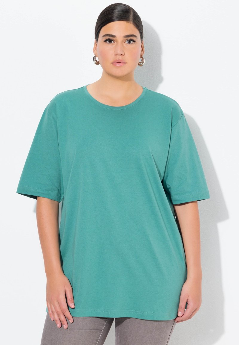 Teal übergroßes T-Shirt, kurze Ärmel, runder Halsausschnitt, weiches Baumwollmaterial. Getragen mit grauen Hosen, die Hände des Models an den Seiten sichtbar.