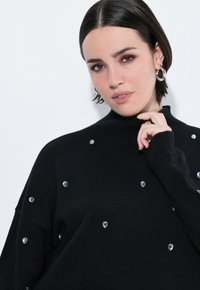 Maglione a collo alto nero con texture a coste, adornato con gemme chiare a forma di cuore sparse. Design aderente con maniche lunghe.