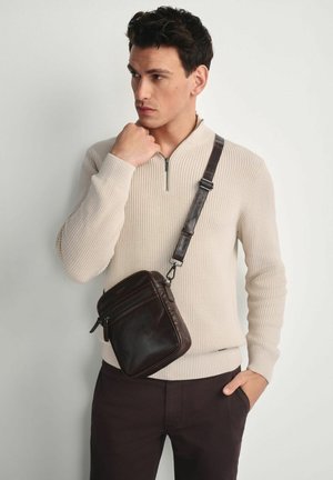 Brązowa skórzana torebka na ramię z regulowanym paskiem, prostokątny kształt, z przednią kieszenią na zamek, noszona przez modelkę w kremowym swetrze w prążki.