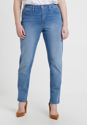 Kvinde iført lyseblå slim-fit jeans og brune højhælede sko, stående mod en ensartet lys baggrund.