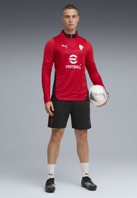 Rød langermet treningstrøye med glidelås og logo, kombinert med svarte shorts. Modellen holder en hvit fotball, iført svarte fotballsko og hvite sokker.