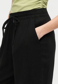 Woman wearing black elastic waist pants with drawstring, hand in pocket, and pale green ruched top visible at waist.