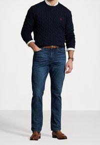 Mörkblå kabelstickad tröja, vit skjorta med krage under, blå jeans, brunt bälte, bruna loafers med gröna prickar, visad i en stående pose.