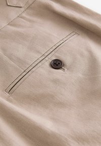 Tan tyg med en slät textur har ett bröstfack och en brun fyra-håls knapp, sydd med matchande tråd.