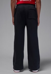 Pantalon de survêtement noir à jambes larges avec une taille élastique, présentant une poche arrière et un tissu doux et texturé. L'ourlet se termine à la cheville.