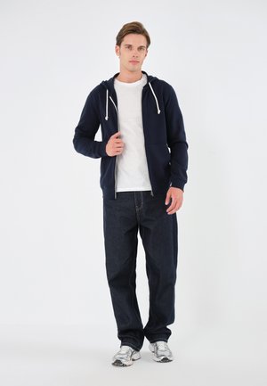 Marinefarbener Zip-Hoodie mit weißen Kordeln, getragen über einem grauen T-Shirt; kombiniert mit dunklen Jeans und silbernen Sneakern.