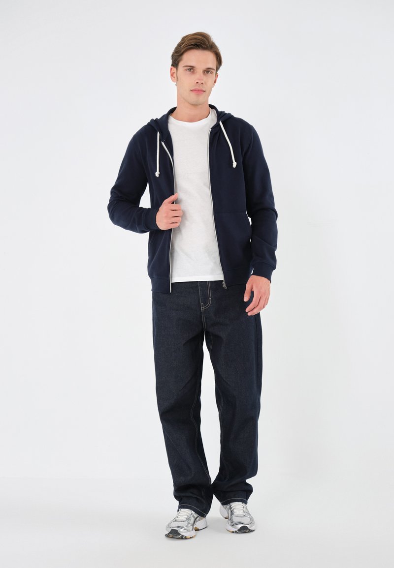 Marinefarbener Zip-Hoodie mit weißen Kordeln, getragen über einem grauen T-Shirt; kombiniert mit dunklen Jeans und silbernen Sneakern.