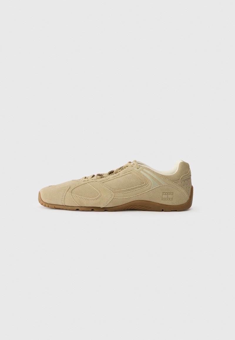 Beige athletic shoe with a textured suede upper, rubber sole, and minimal design accents. Features elastic laces and subtle logo detailing.