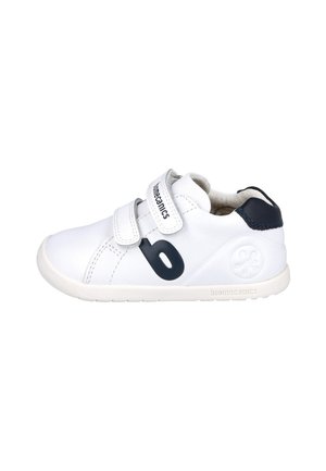 Zapato de cuero blanco con acentos en azul marino, dos correas de velcro, punta redondeada y suela texturizada. Presenta un logo en el lateral.