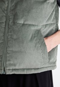 Vesta din catifea verde cu textură ribbed verticală, închidere cu fermoar și buzunare laterale. Marginile de la partea de jos sunt finisate cu un tiv întors.