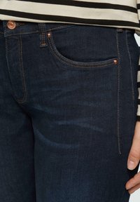 Dunkelblaue Denim-Jeans mit Kupfernieten, Kontrastnähten und einer vorderen Tasche. Glatte Textur, schlanke Passform und schmal zulaufendes Bein.