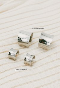 Sudraba apli auskari divos izmēros (S un L). Gluda metāla apdare ar izliektiem formām, novietoti uz teksturēta krēmkrāsas auduma fona.