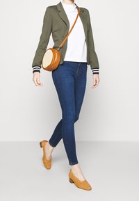 Blazer verde oliva con polsini a righe, dolcevita bianca, jeans skinny blu scuro e scarpe con tacco block in suede beige; borsa rotonda.