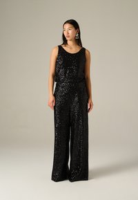 Jumpsuit noir en sequins avec design sans manches, pantalons évasés et encolure ronde. Texture lisse et surface brillante avec une taille cintrée.