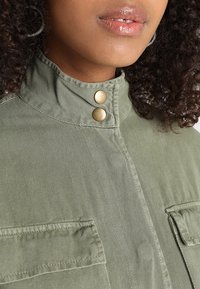 Olive green collar jacket featuring two gold snap buttons, textured fabric, and two front pockets with stitched detailing.